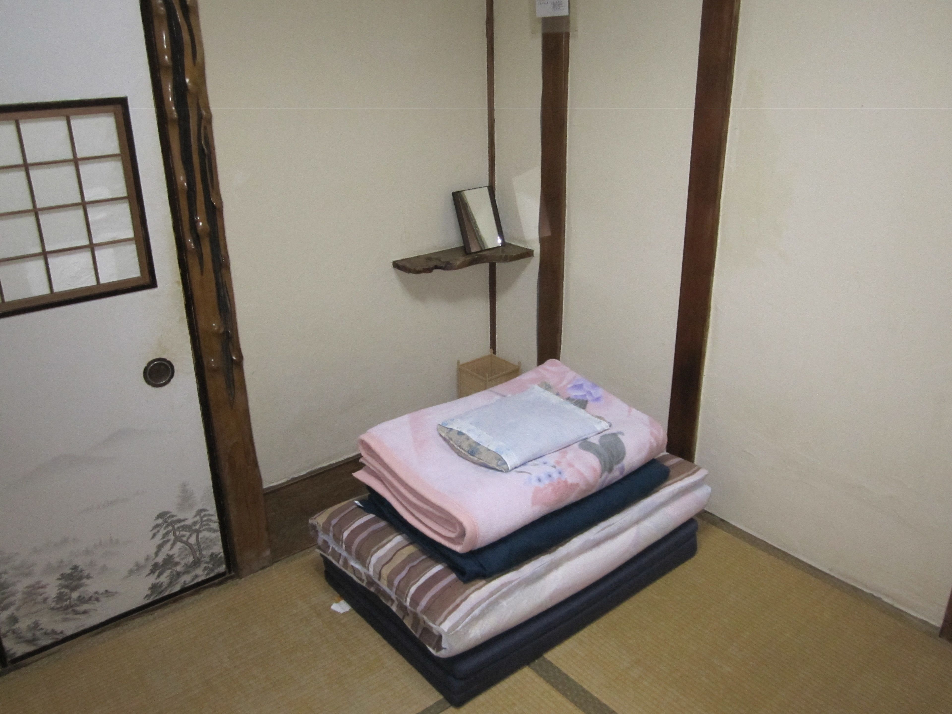 A room with a bed, a wooden door with a grid pattern, a small shelf, and a wall with a traditional design.