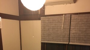 A traditional Japanese-style room with tatami flooring, a low wooden bed with a pillow, a small table with a remote control, and a window with a blind. - Taito Ryokan (Tokyo)
