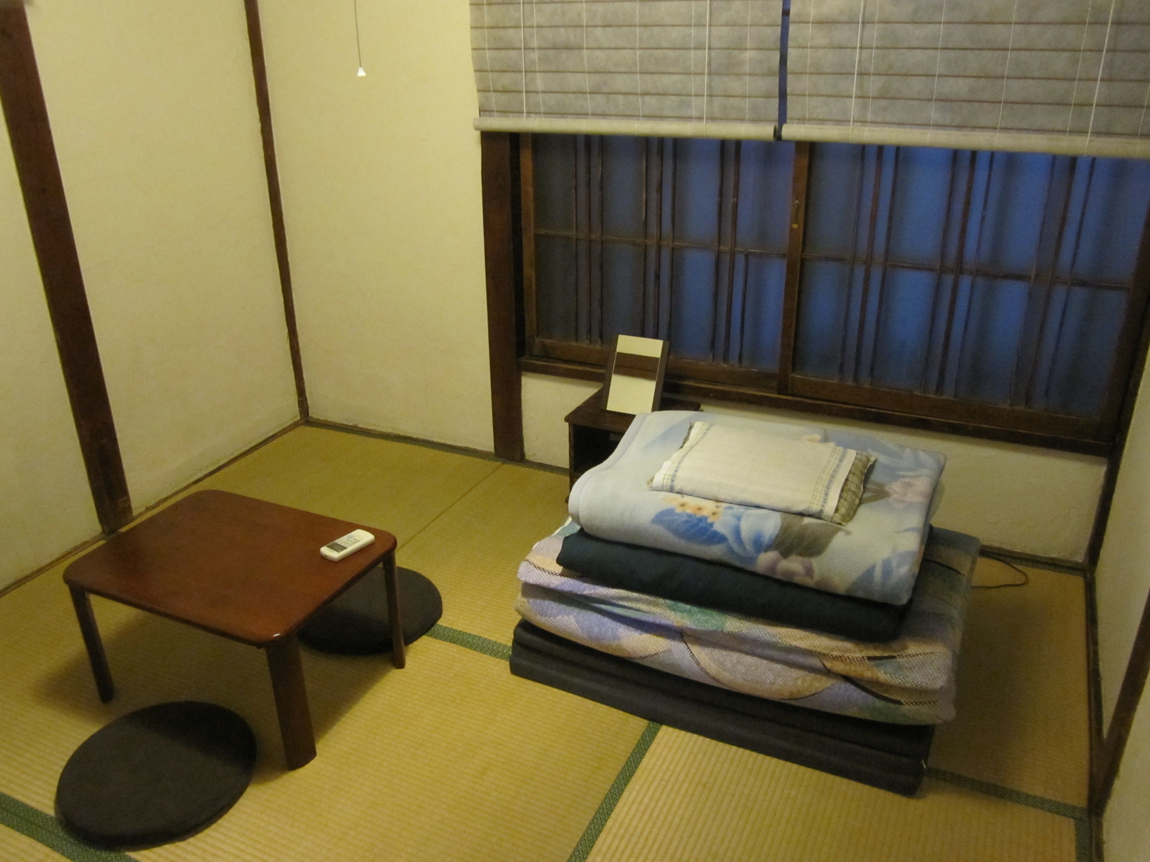 A traditional Japanese room with tatami flooring, a low wooden table, a futon bed, and a window with blue curtains.