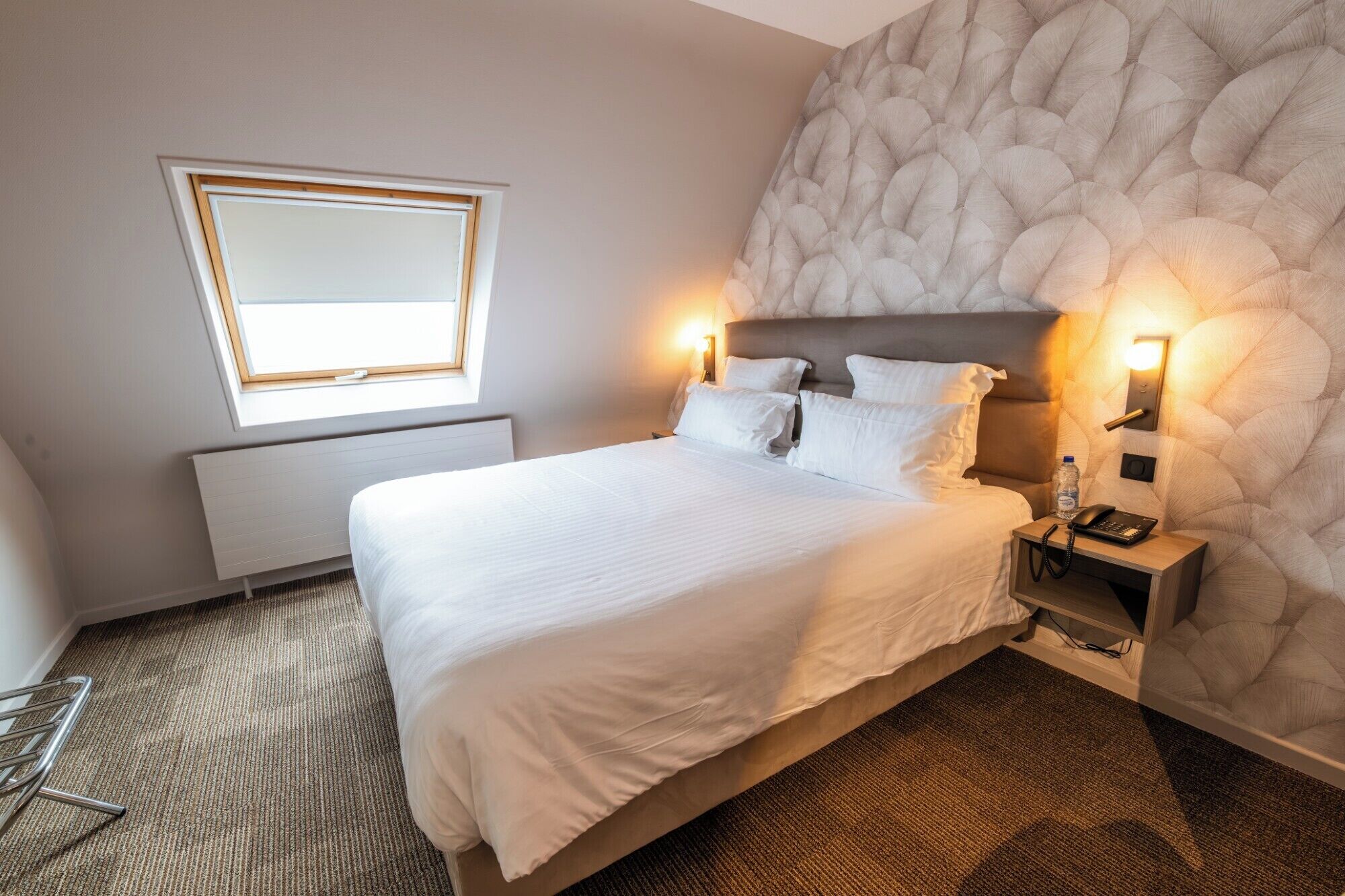 A hotel room with a bed, bedside tables, a wall-mounted light, a radiator, a window, and a chair.