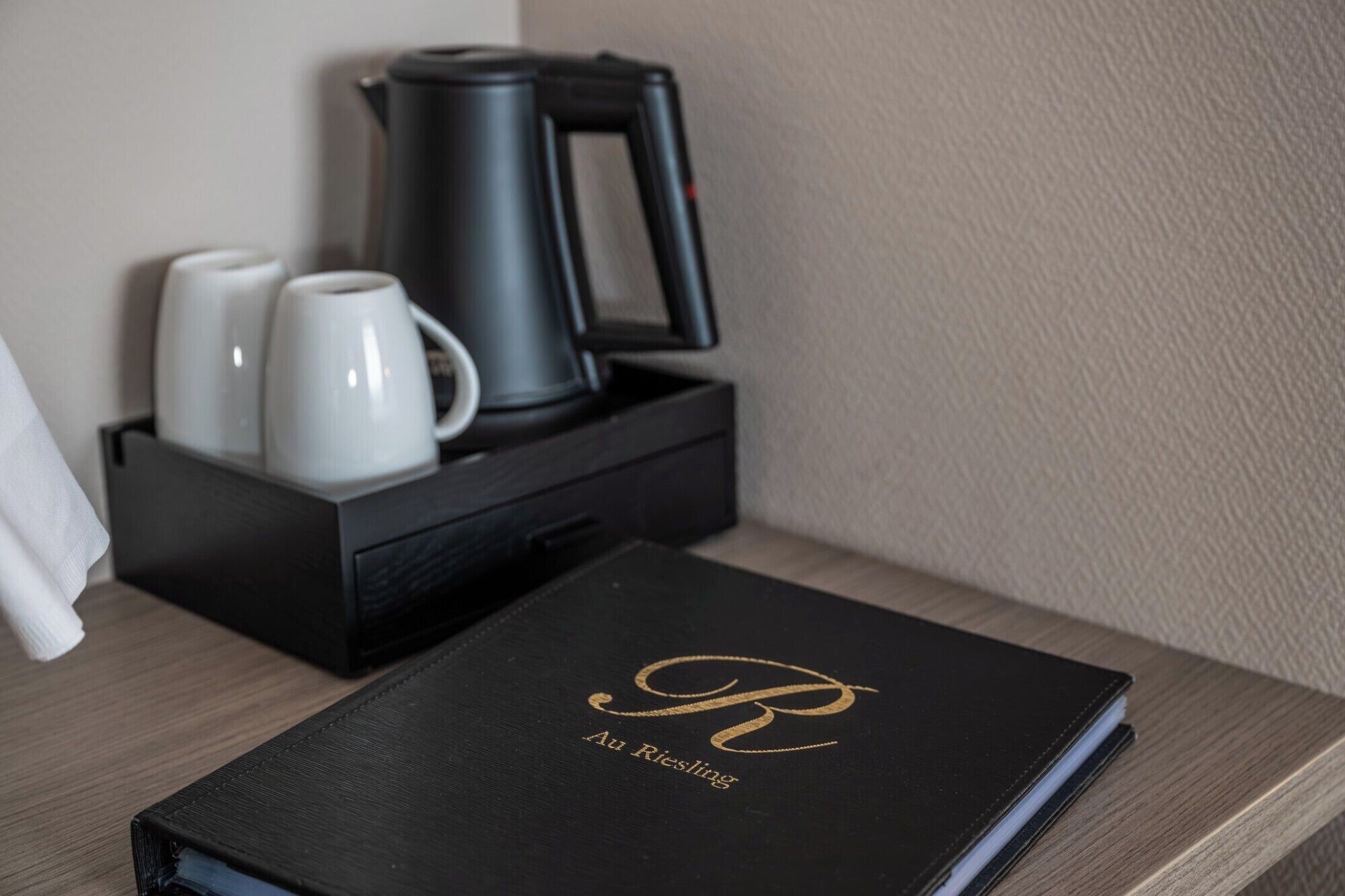 Een zwart lederen boek met gouden letters op een houten oppervlak, een zwarte dienblad met een koffiezetapparaat en twee witte mokken, en een witte zakdoos.