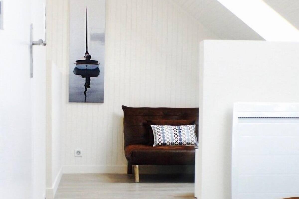 Une pièce avec un plancher en bois, un canapé marron agrémenté de coussins à motifs, un climatiseur blanc et une œuvre d’art représentant un bateau, accrochée au mur.