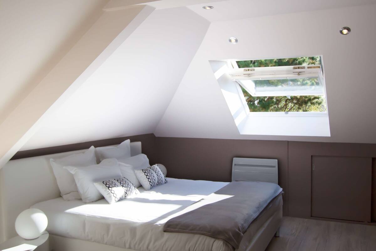 Une chambre moderne avec un plafond en pente, une lucarne, un lit avec une literie blanche et une table de chevet avec une lampe.