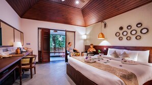 A hotel room with a wooden ceiling, a large bed with white linens, a desk with a lamp, and a view of greenery through an open door. - The Elephant Court (Peermade)