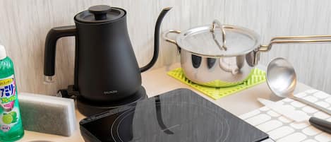 A kitchen with a black electric kettle, a stainless steel pot on a green mat, a green bottle of dish soap, a white sponge, and a spoon.