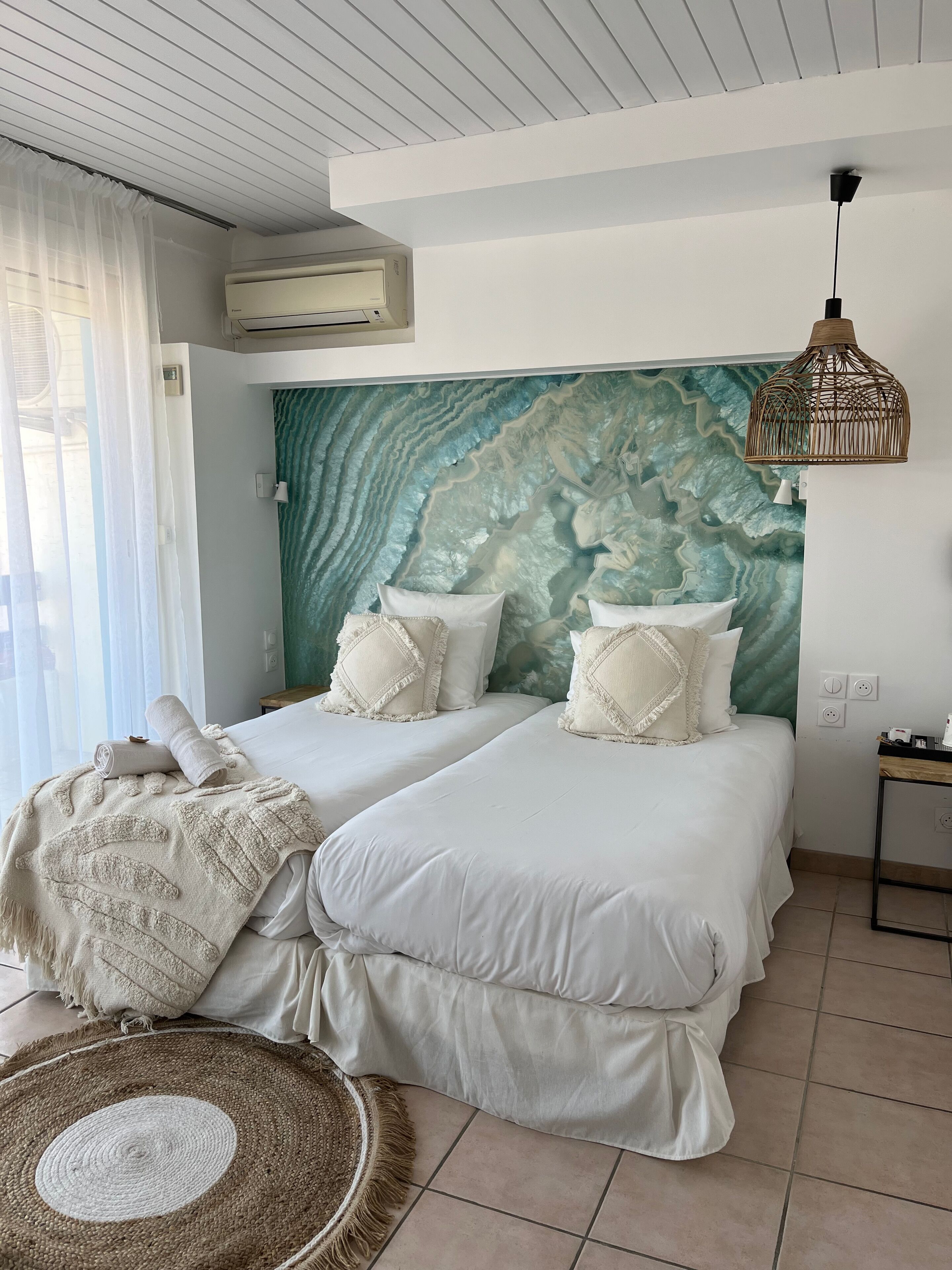 Une chambre d’hôtel avec deux lits, une grande fresque murale à motifs, un fauteuil en rotin tressé et un ventilateur de plafond.