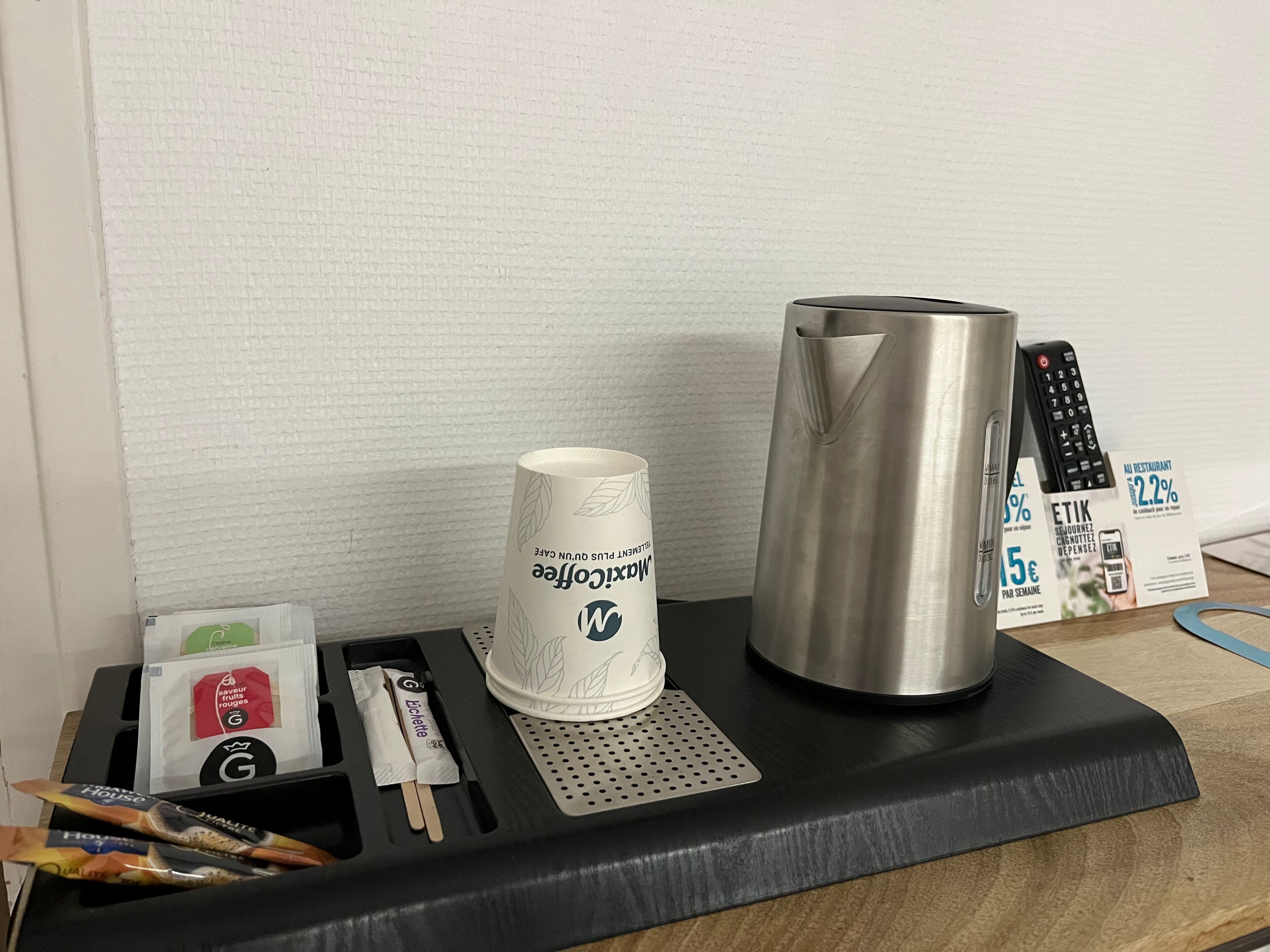 A hotel minibar with a coffee cup, a kettle, a remote control, and some toiletries.