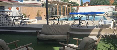 A rooftop pool area with outdoor seating, including lounge chairs and a table, and a view of the surrounding buildings.