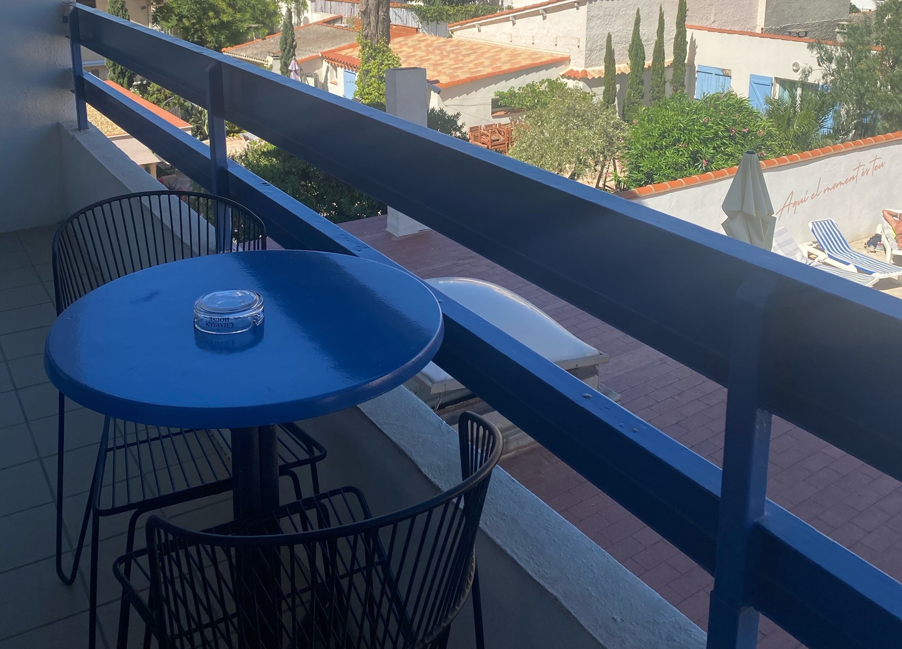Un balcon avec une table et des chaises bleues, donnant sur une cour intérieure bordée d’arbres et entourée de bâtiments.