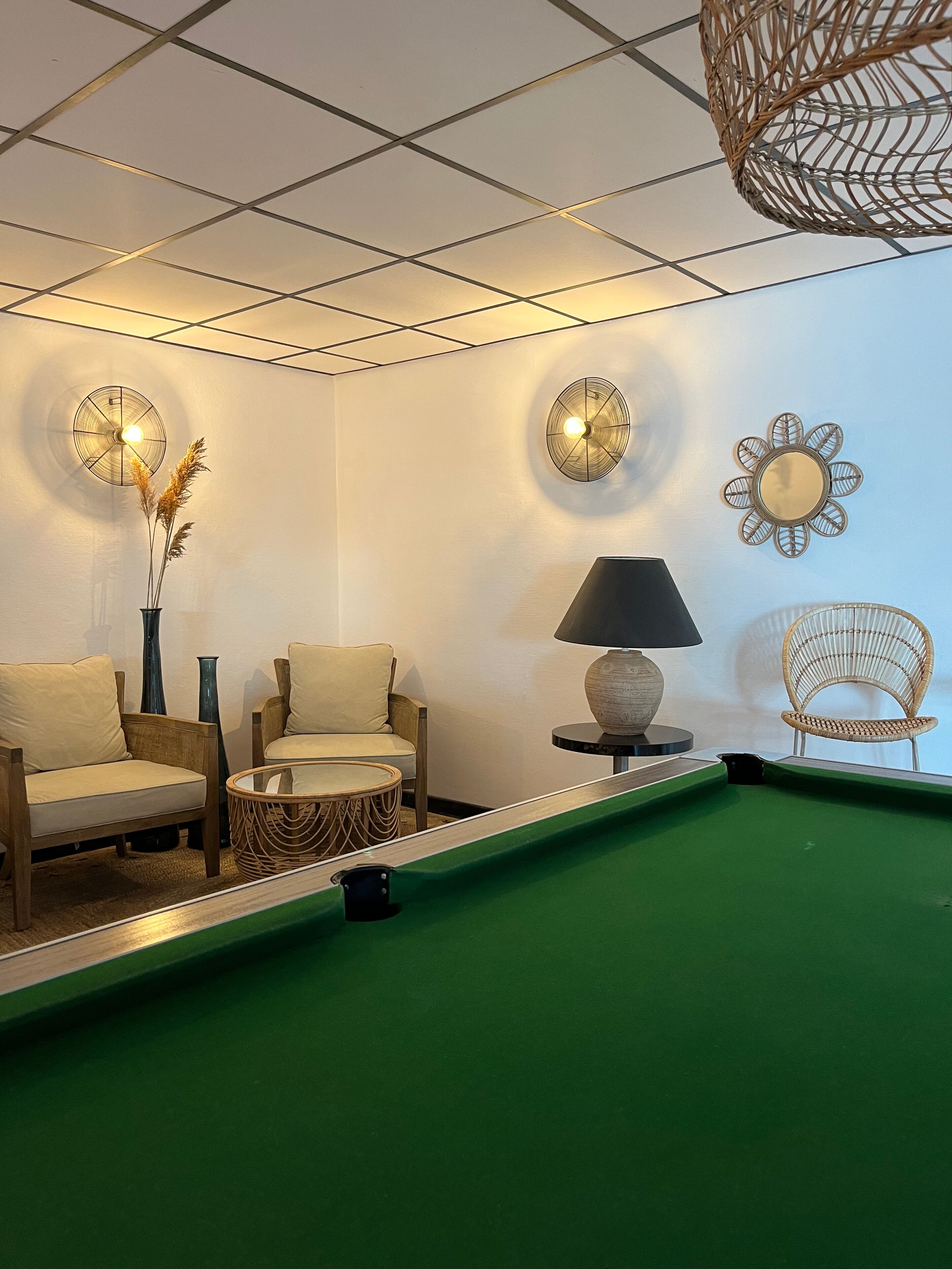 A pool table with a green felt surface, surrounded by two beige sofas and a wicker chair. There are two wall-mounted lamps with round bases and flower-like designs.