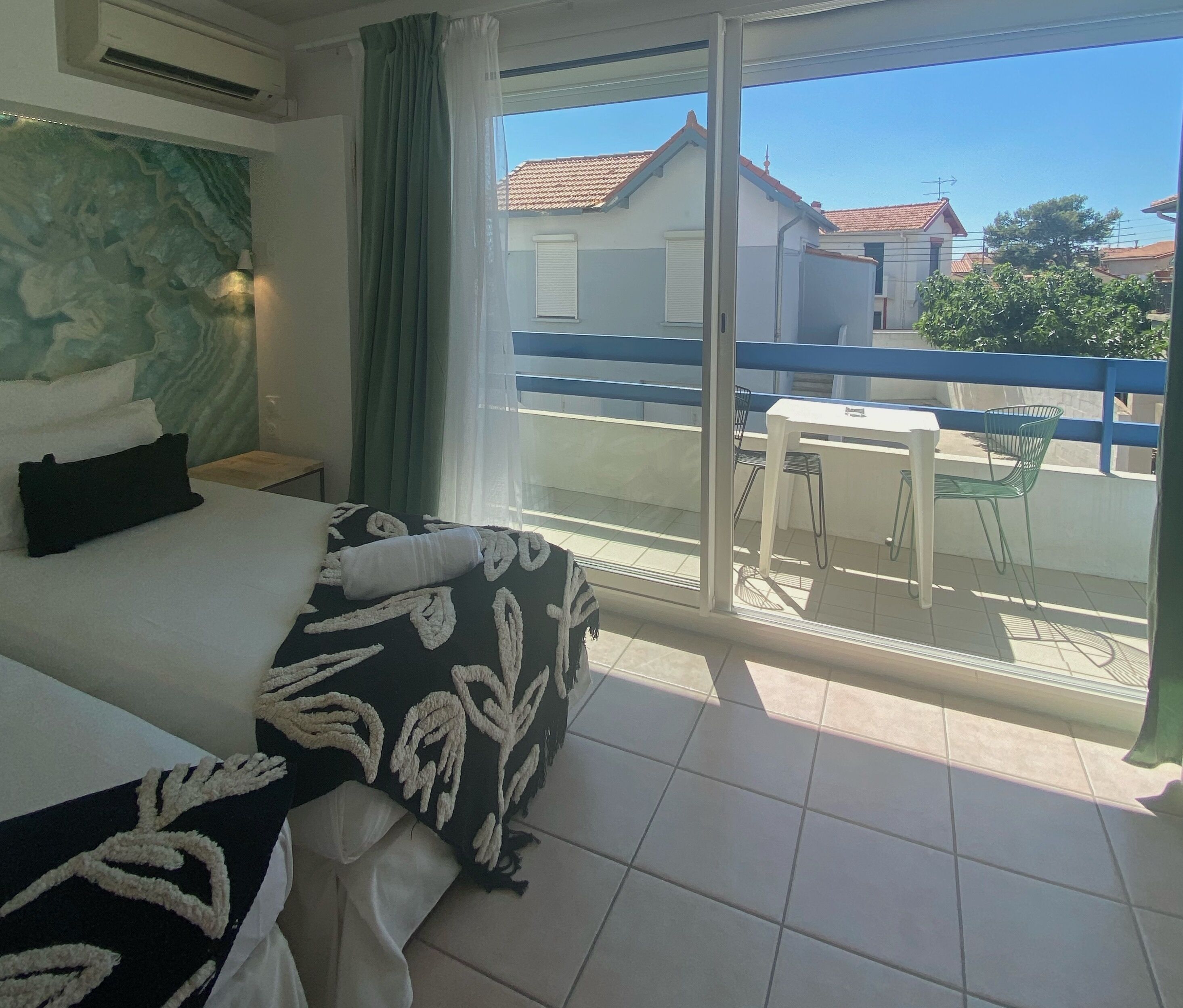 A hotel room with a bed, a small table, and a balcony with a view of the neighborhood.