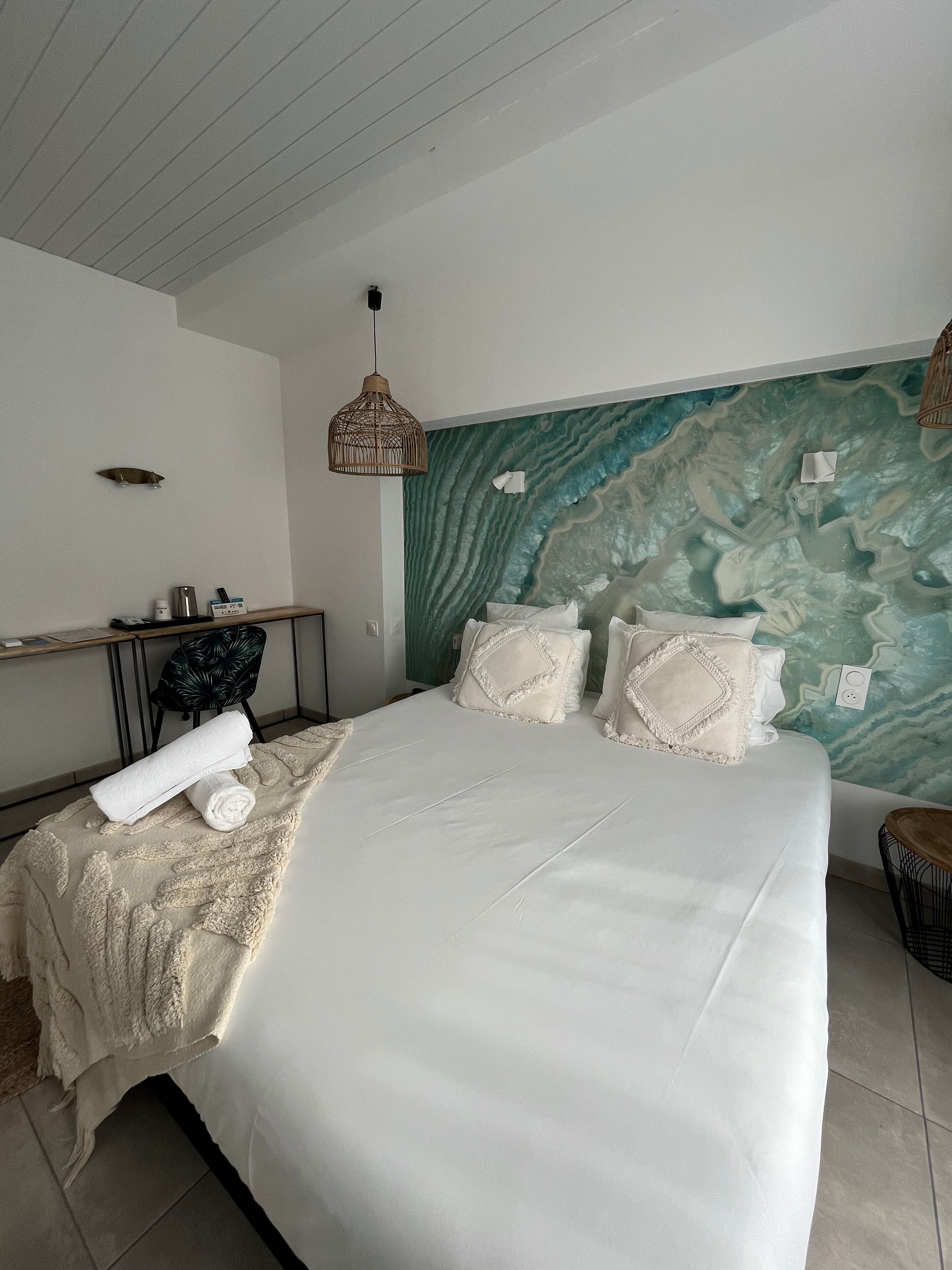 En pænt redt seng med hvide sengetøj og dekorative puder, et træsidet bord med en lampe, en stol og en væg med et struktureret muralmaleri.