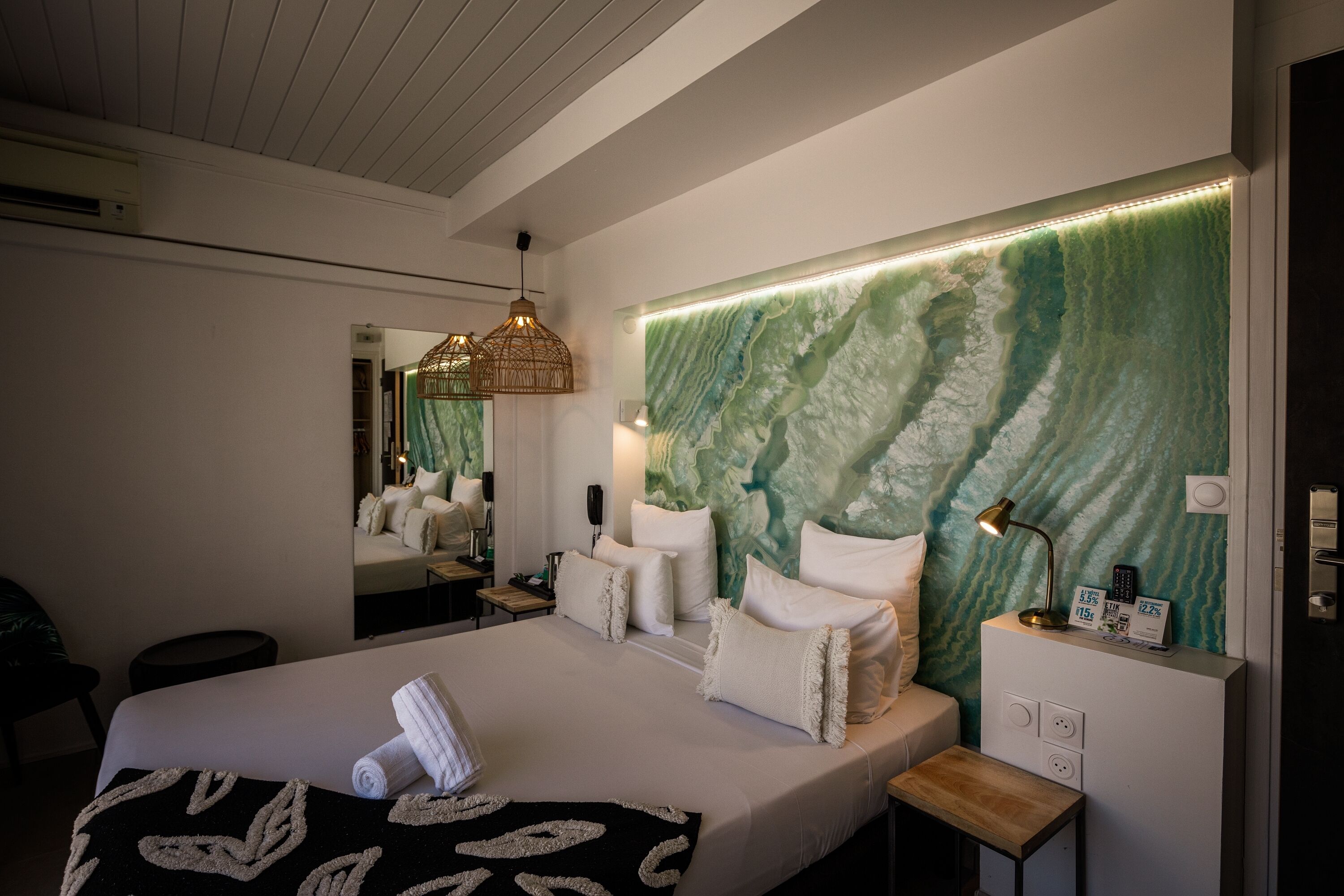 Une chambre d’hôtel moderne avec un grand lit, une table de chevet avec une lampe, un miroir fixé au mur et un mur texturé de couleur verte.