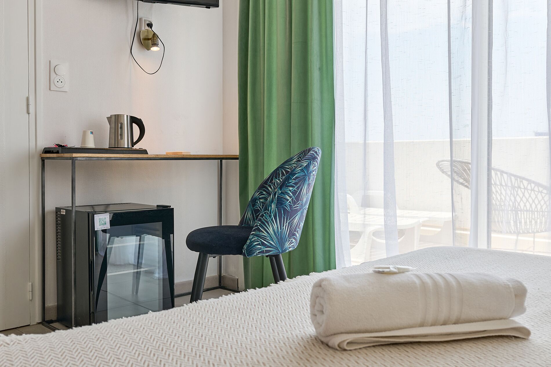 A hotel room with a bed, a desk with a computer, a chair, and a view of the balcony.