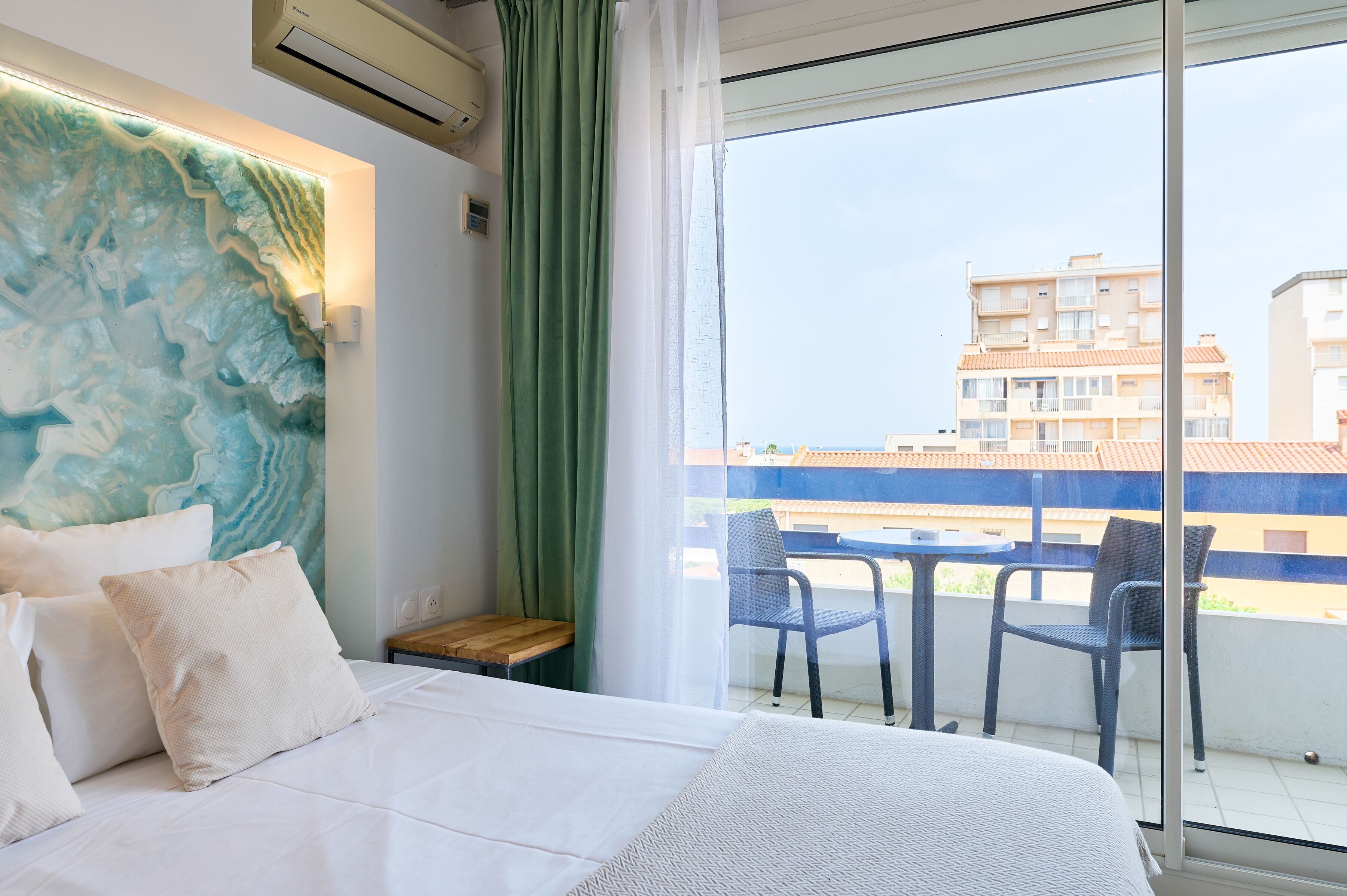 Habitació d'hotel amb un llit, una cortina verda, una vista de la ciutat a través d'una finestra gran i un balcó amb cadires.