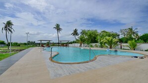 A swimming pool with a curved design, surrounded by a paved walkway and palm trees. - Hotels Diamond Glory - Mandarmani (Kanthi)
