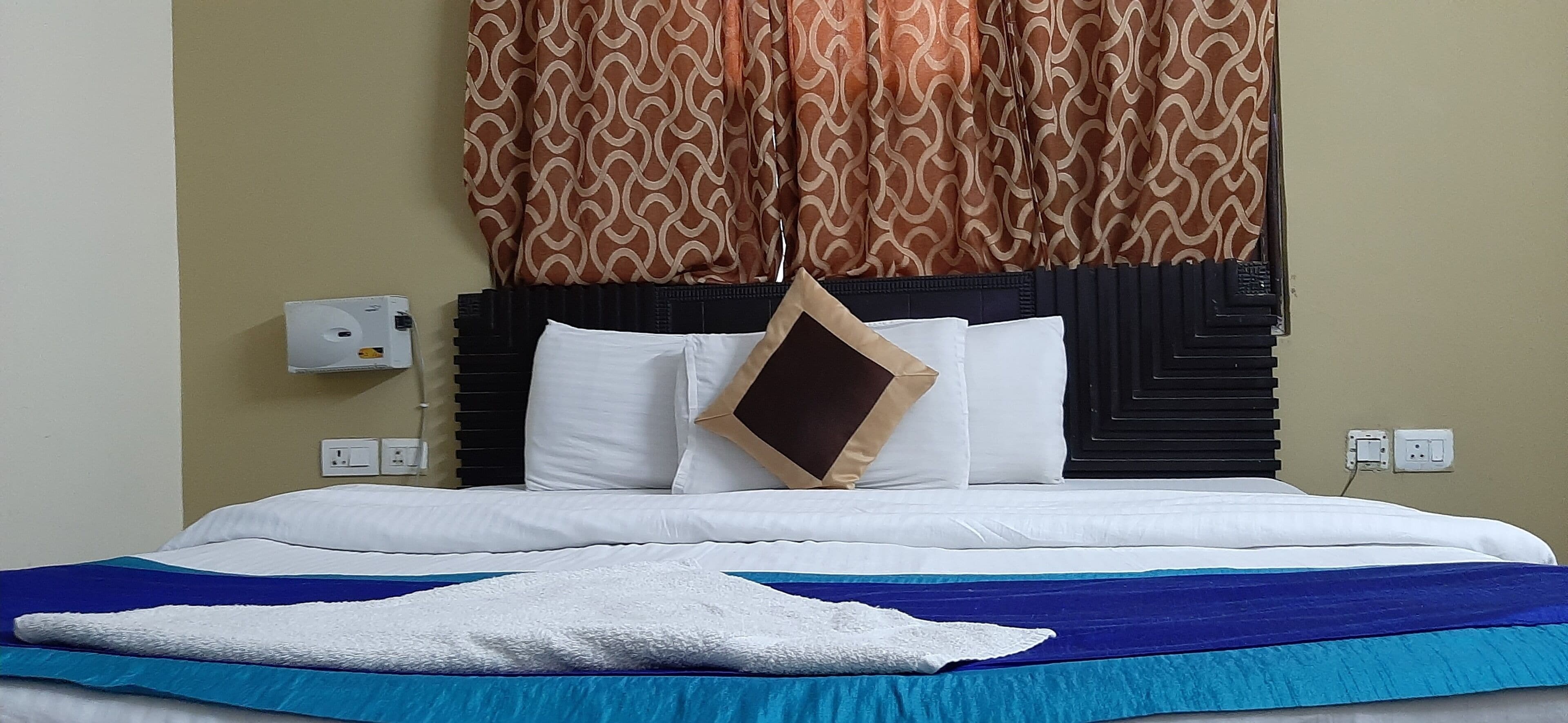 A hotel room with a bed, a white pillow, a brown pillow, a blue blanket, and a brown accent pillow.