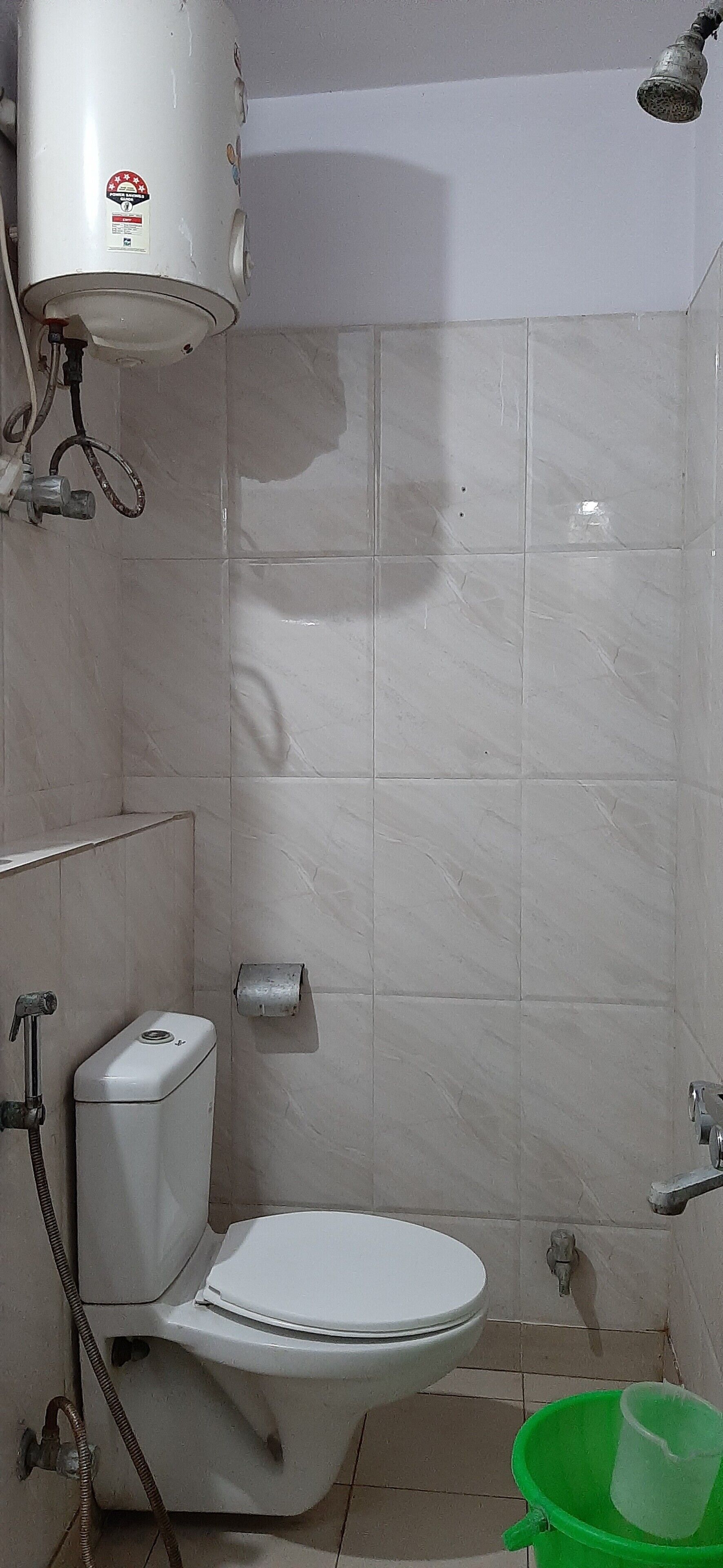 A bathroom with a white toilet, a green bucket, and a water heater mounted on the wall.