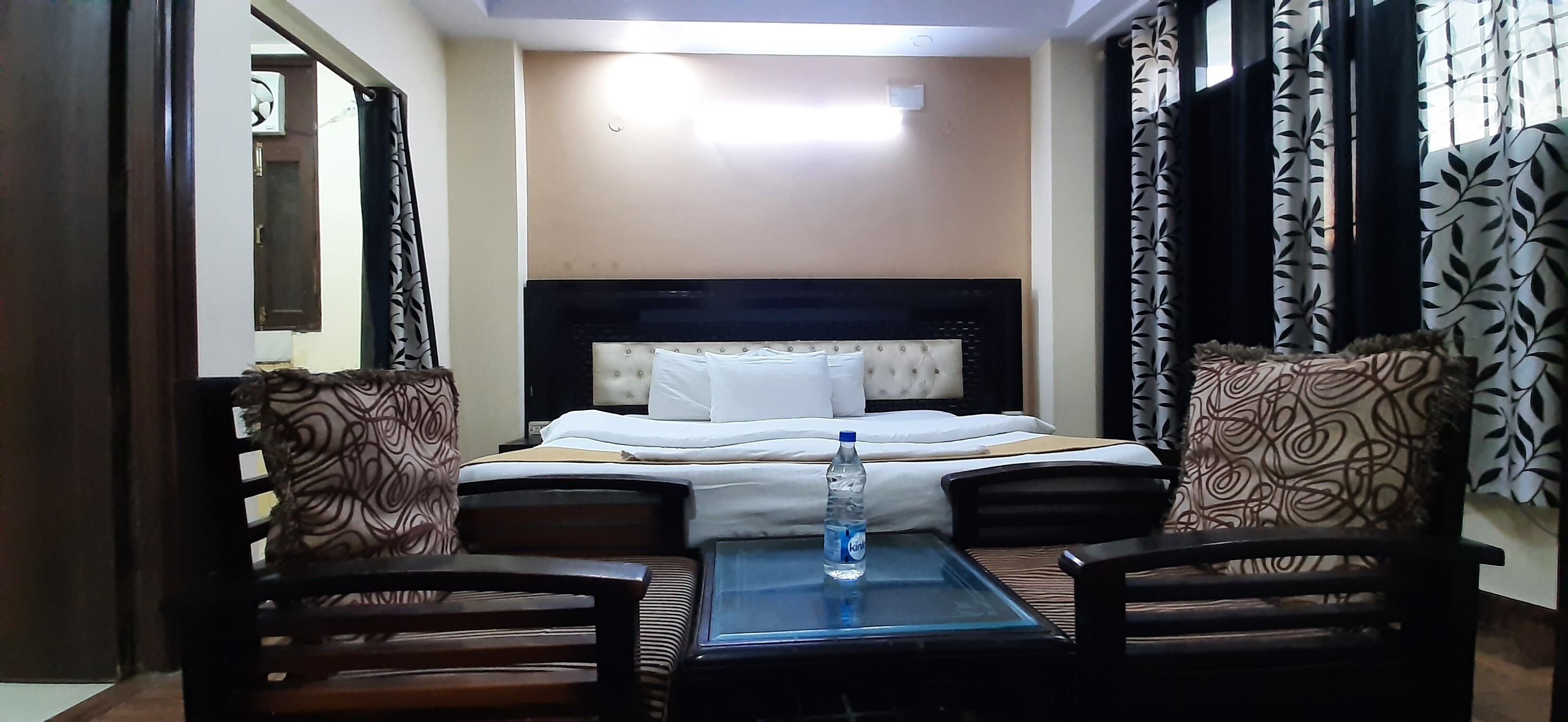 A hotel room with a bed, two patterned armchairs, a small table with a water bottle, and curtains with a leaf pattern.