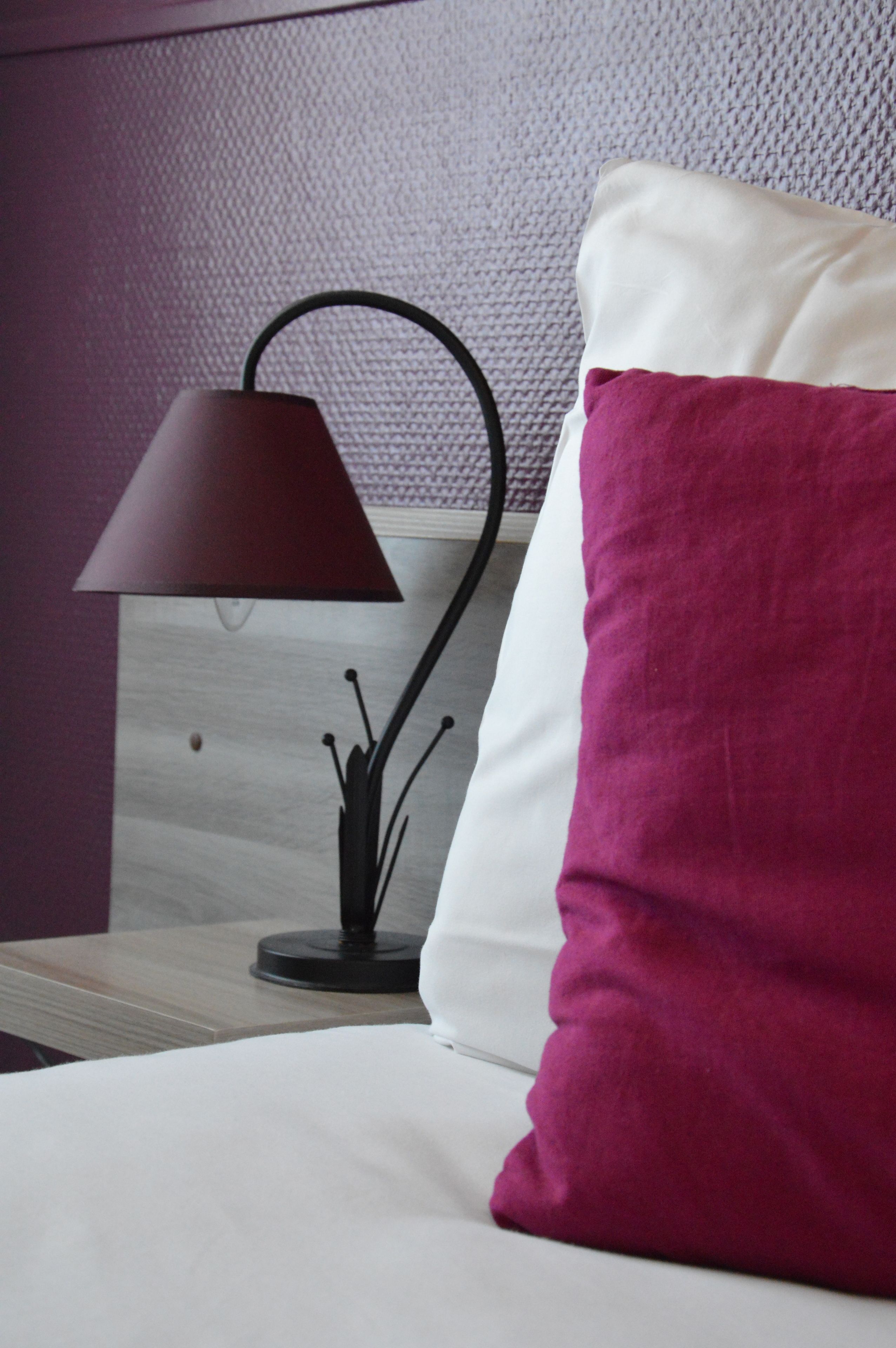 A neatly made bed with a white and purple pillow, a black lamp with a round base, and a textured wall behind.