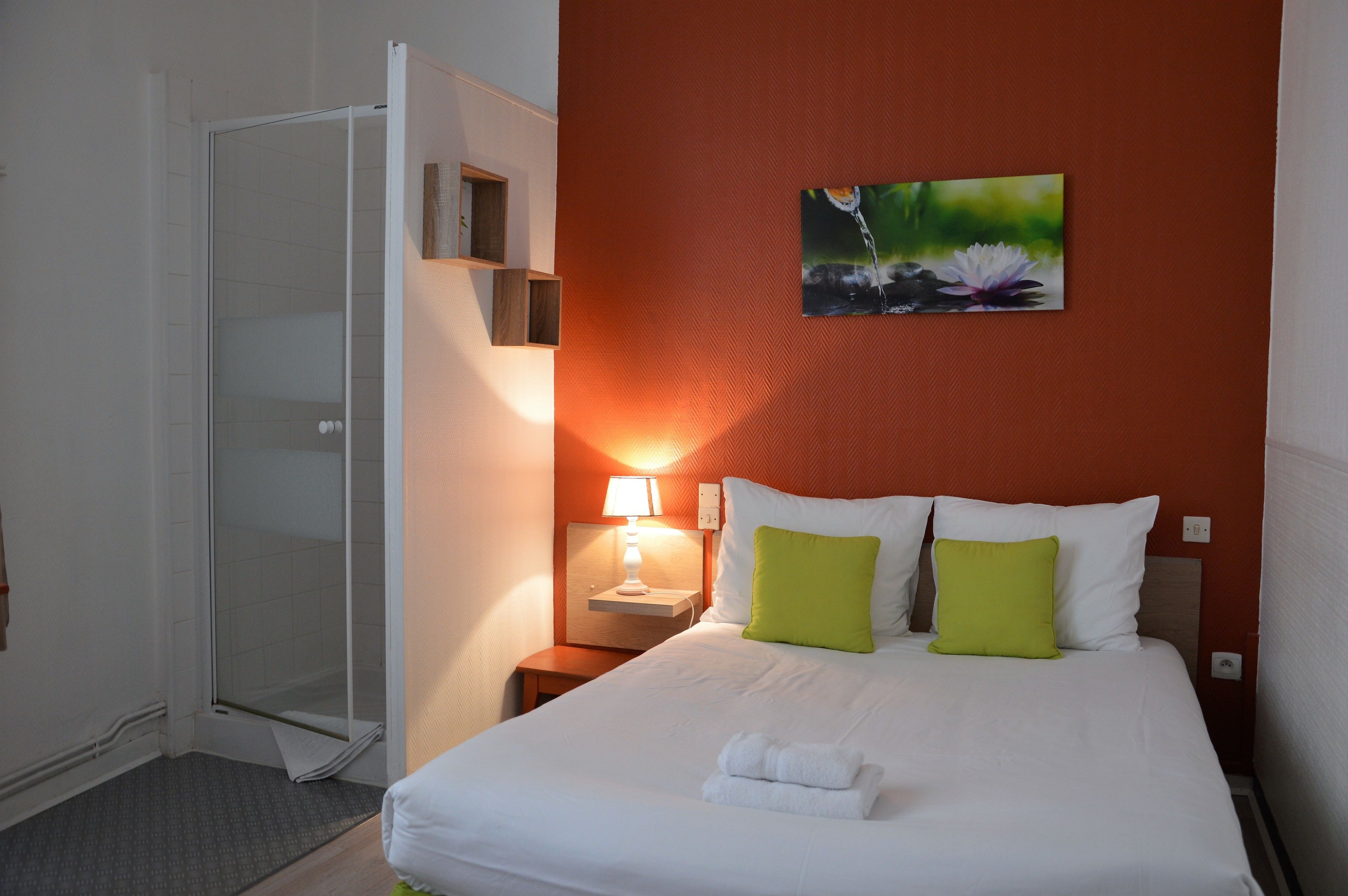 Habitación de hotel con cama, mesita de noche con lámpara, un cuadro en la pared y puerta corredera de cristal que da a la zona de ducha.