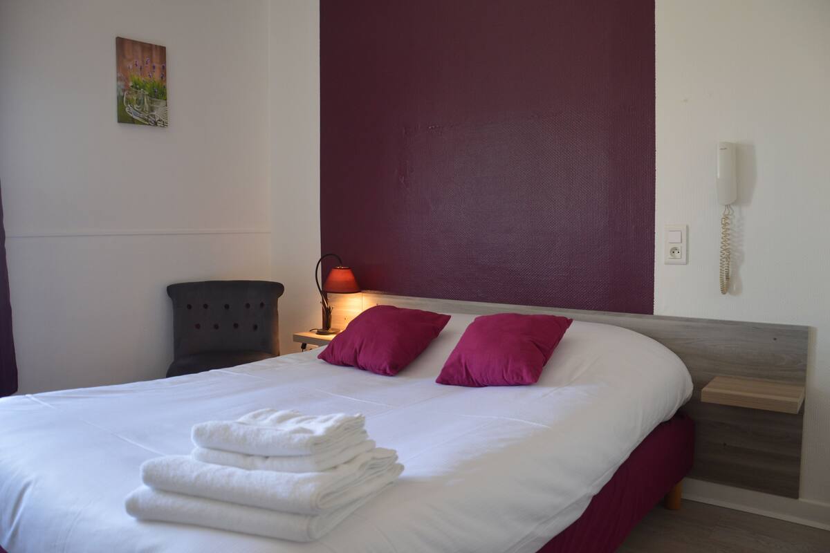 Een net opgemaakt bed met witte lakens en twee paarse kussens, een bedlampje, een telefoon en een klein kunstwerk aan de muur.