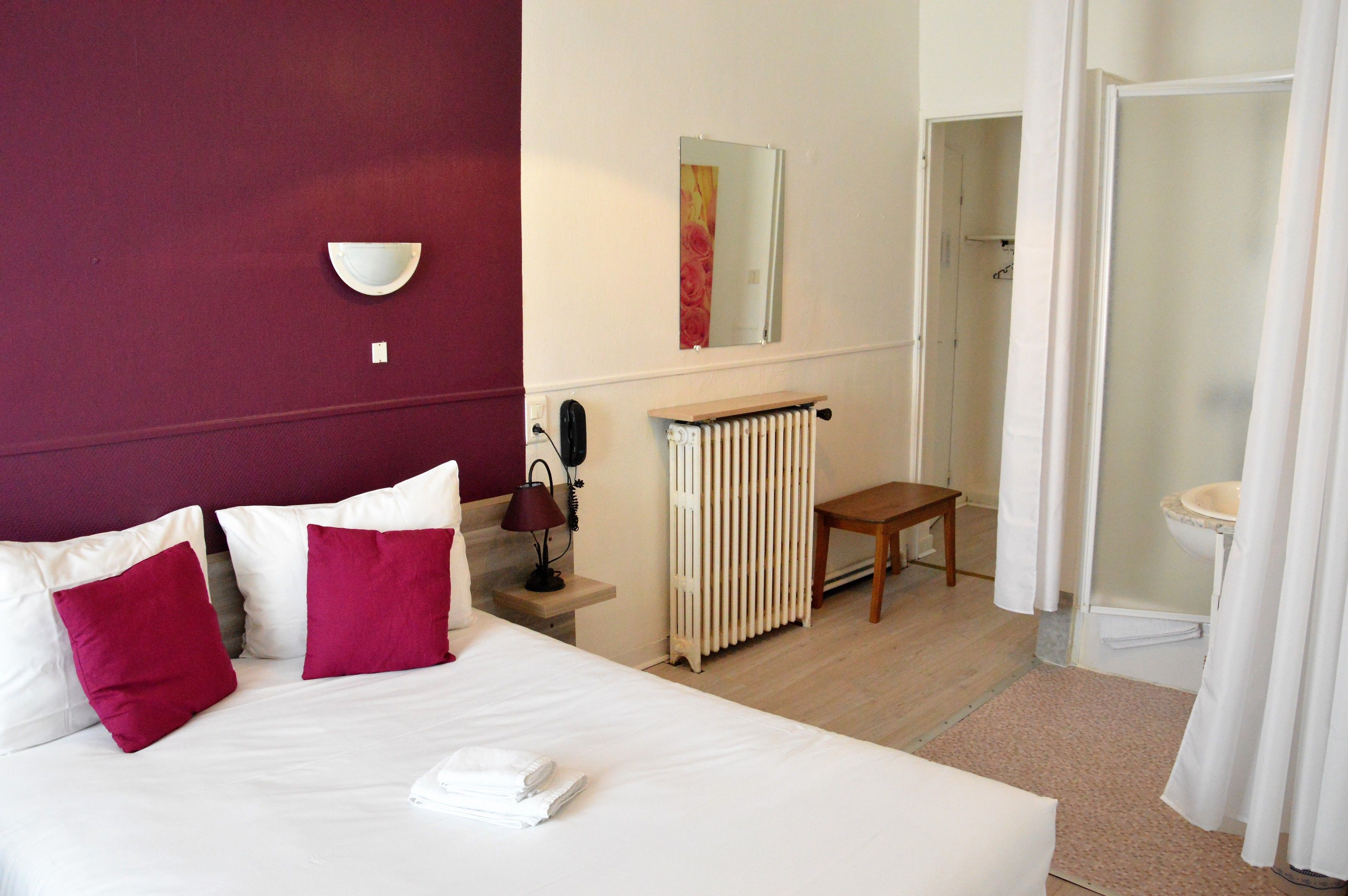A hotel room with a bed, a bedside table with a lamp, a radiator, a small wooden stool, and a bathroom visible through an open door.