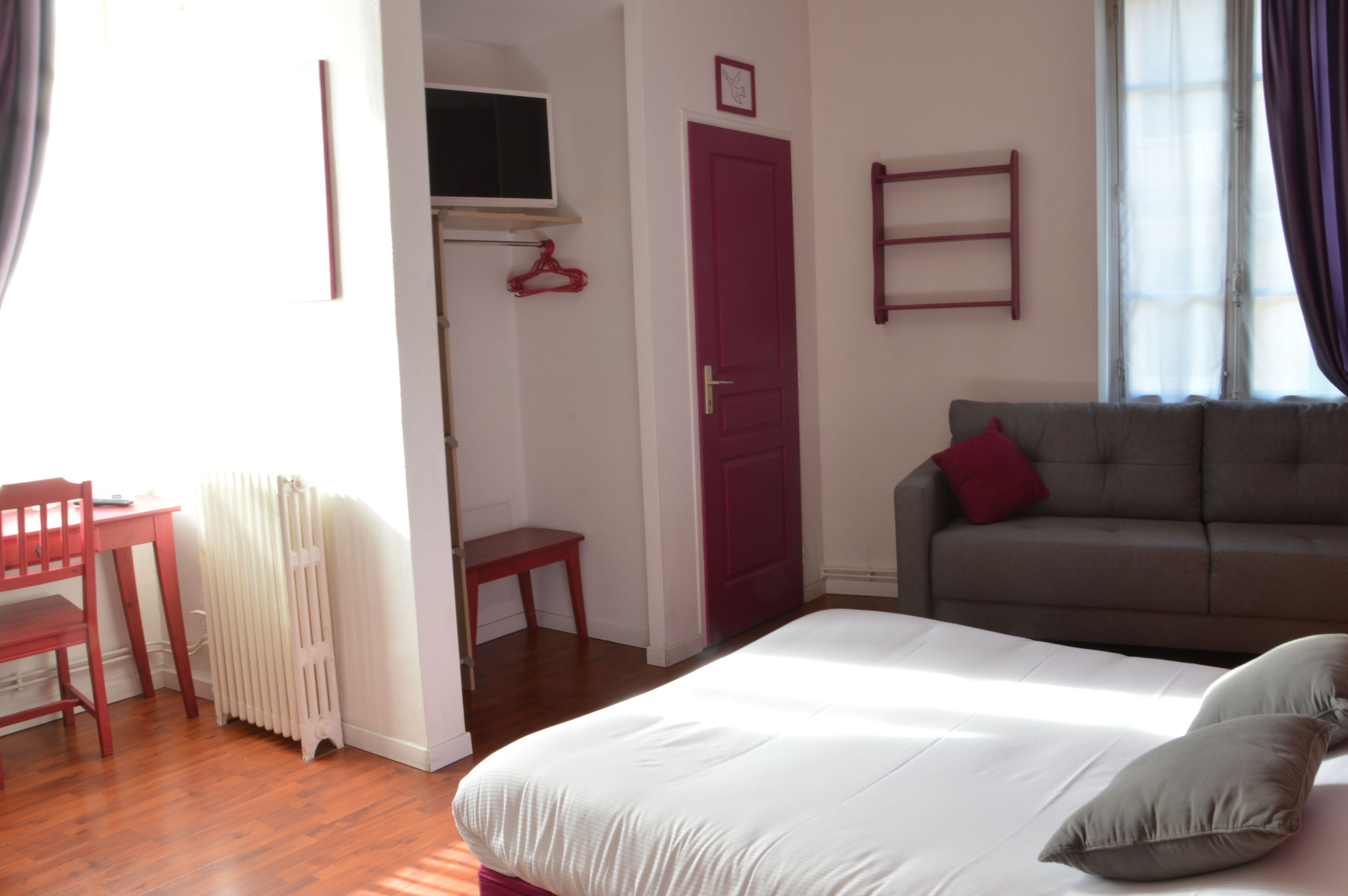 Une chambre à coucher avec un lit, une porte rouge, un canapé gris et une fenêtre avec des rideaux.