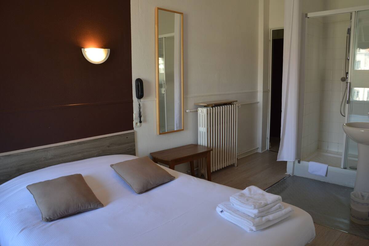 Hotelkamer met een bed, nachtkastje, spiegel, radiator en een badkamer met wastafel en douche.