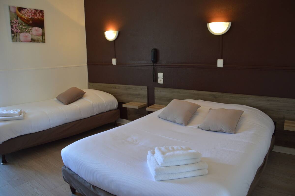 Een hotelkamer met twee bedden, elk met een kussen en een opgevouwen handdoek. Er hangen wandlampen boven de bedden en aan de muur zit een telefoon.