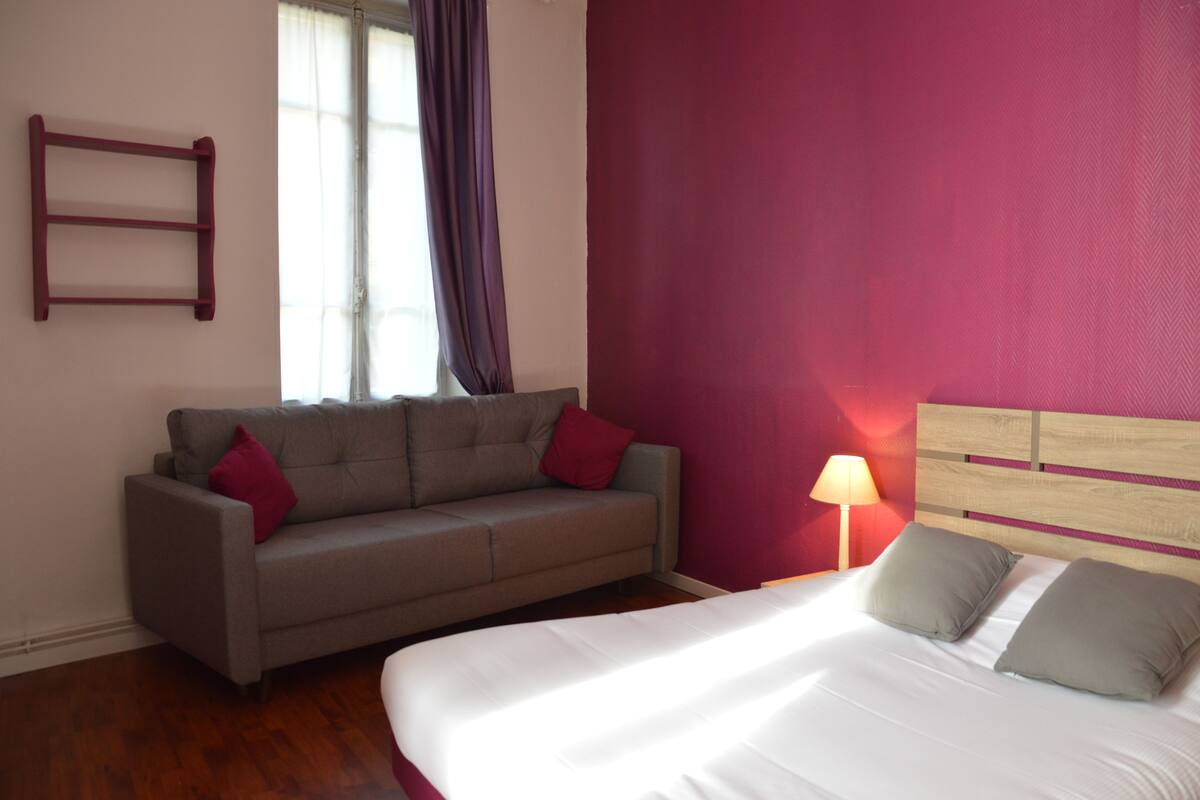 Een slaapkamer met een bed, een grijze bank met rode kussens, een houten hoofdbord, een raam met gordijnen en een wandplank.