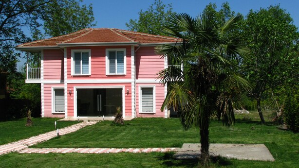 A two-story pink house with a red roof, a front door, and a small front yard with a palm tree and a paved path leading to the entrance. - Botanika Hotel Sapanca (Sapanca)