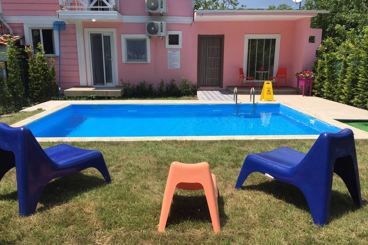 Una piscina con dos sillas azules y una silla naranja en primer plano, rodeada de un césped y un edificio rosa con ventanas y equipos de aire acondicionado.