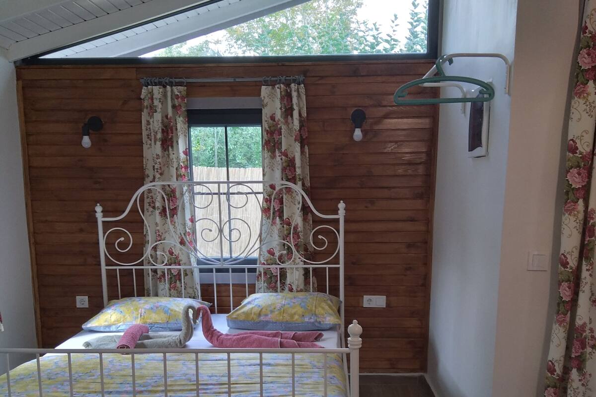 Un dormitorio con pared de madera, una ventana con cortinas, una cama con ropa de cama floral y una lámpara colgante.