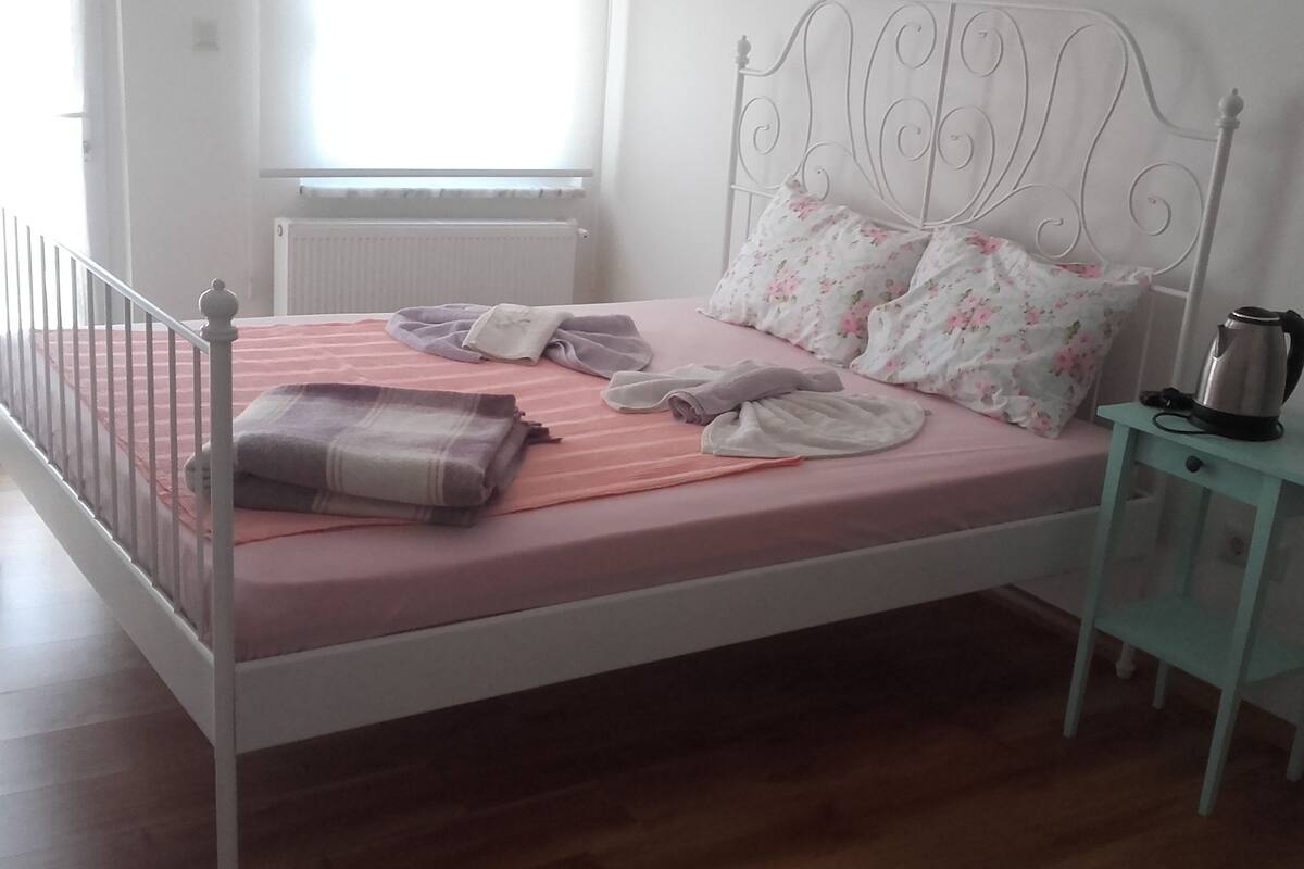Una cama bien hecha con ropa de cama rosa, cabecero de metal y una mesita de noche con hervidor.