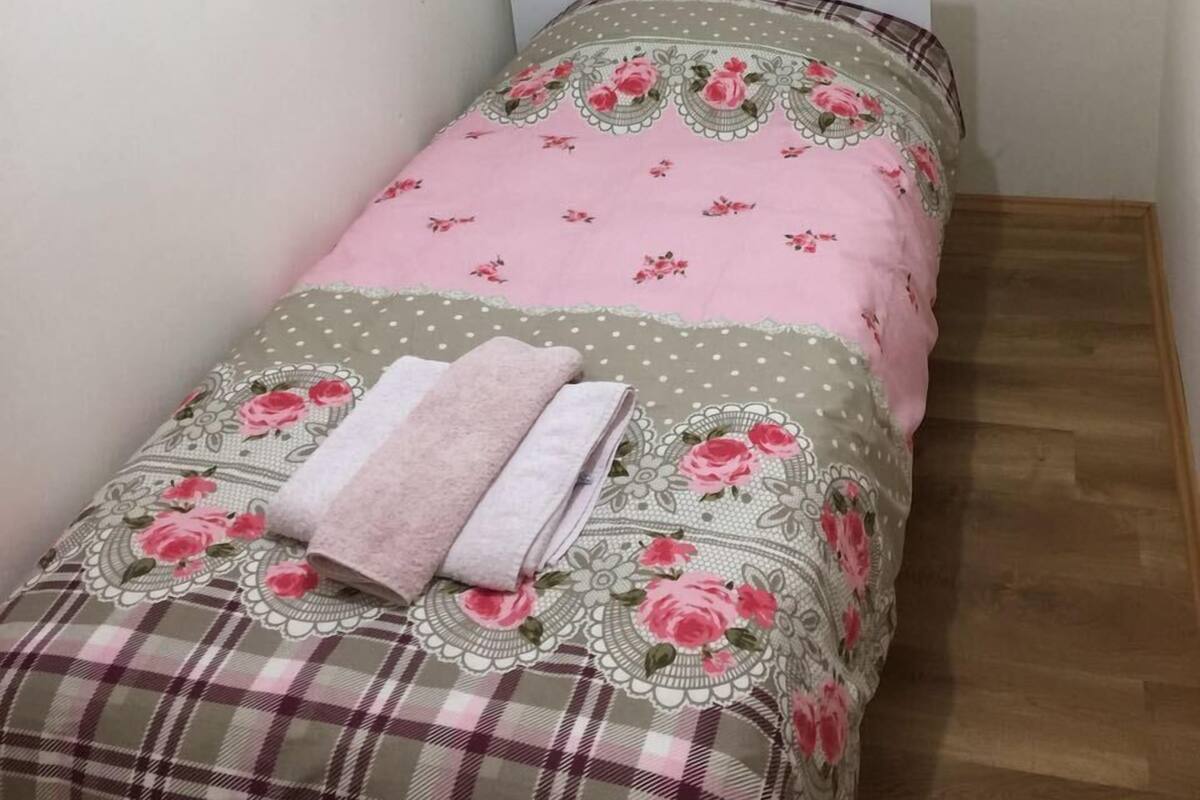 Una cama pequeña y ordenada con ropa de cama floral y detalles a cuadros, una lámpara de pie y una ventana con persianas.