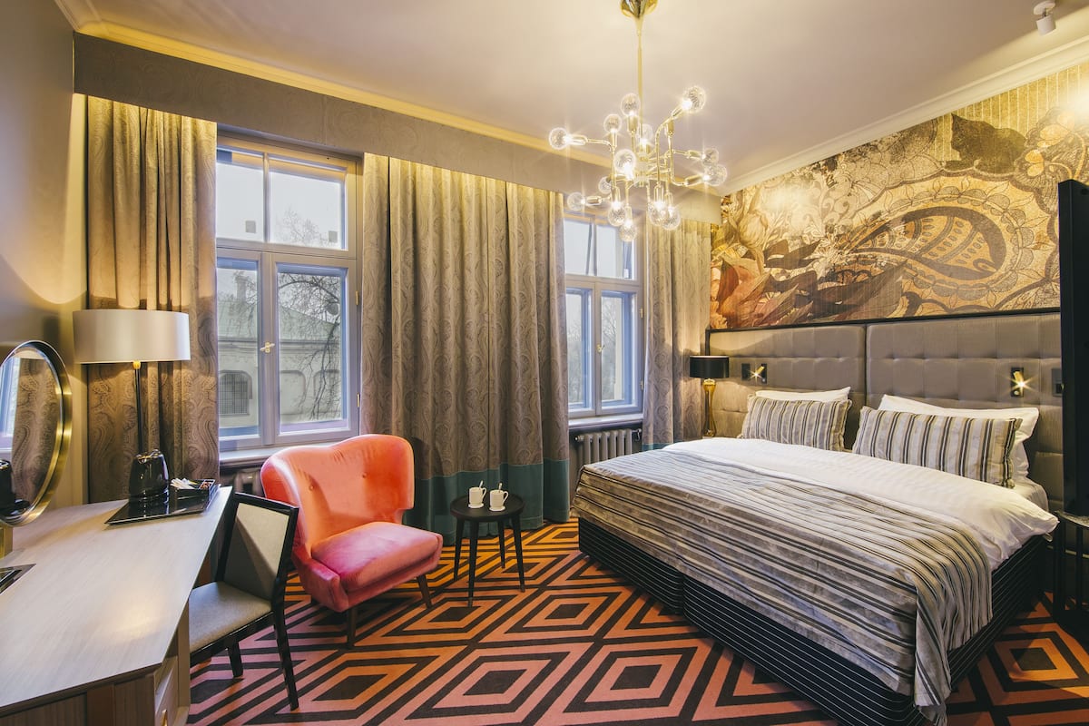 A hotel room with a large bed, a desk with a chair, a small table with two cups, and a large window with curtains.