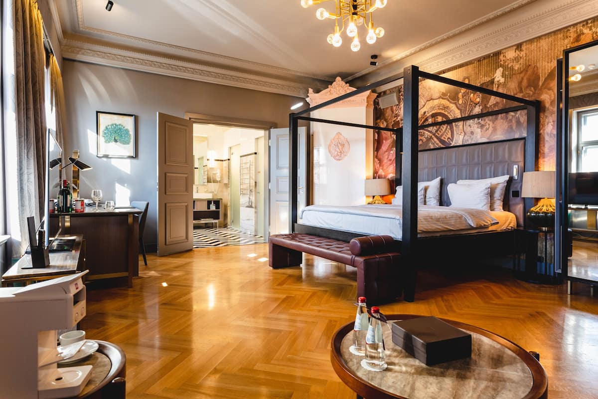 A spacious hotel room with a four-poster bed, a sitting area with a coffee table, and a view of the hallway.