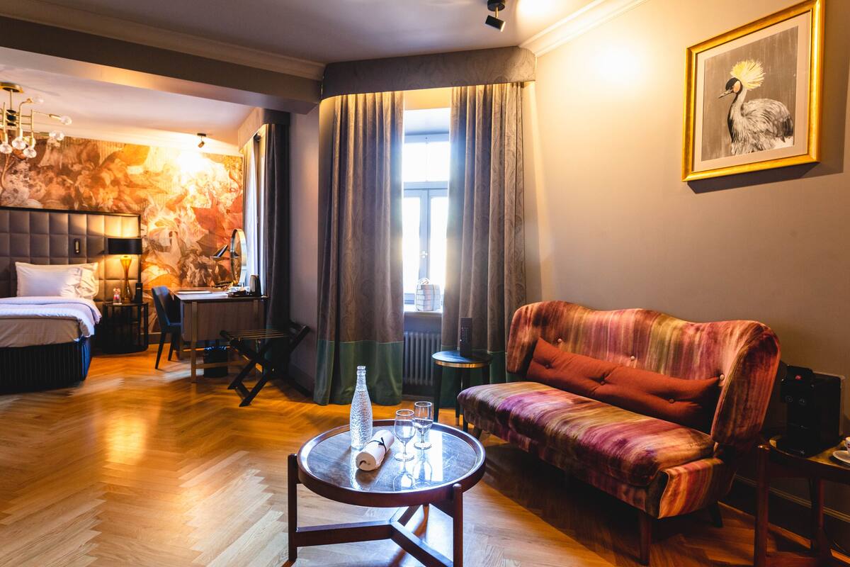 A hotel room with a bed, a sofa, a round table with a bottle and glasses, a chair, a desk, and a painting on the wall.