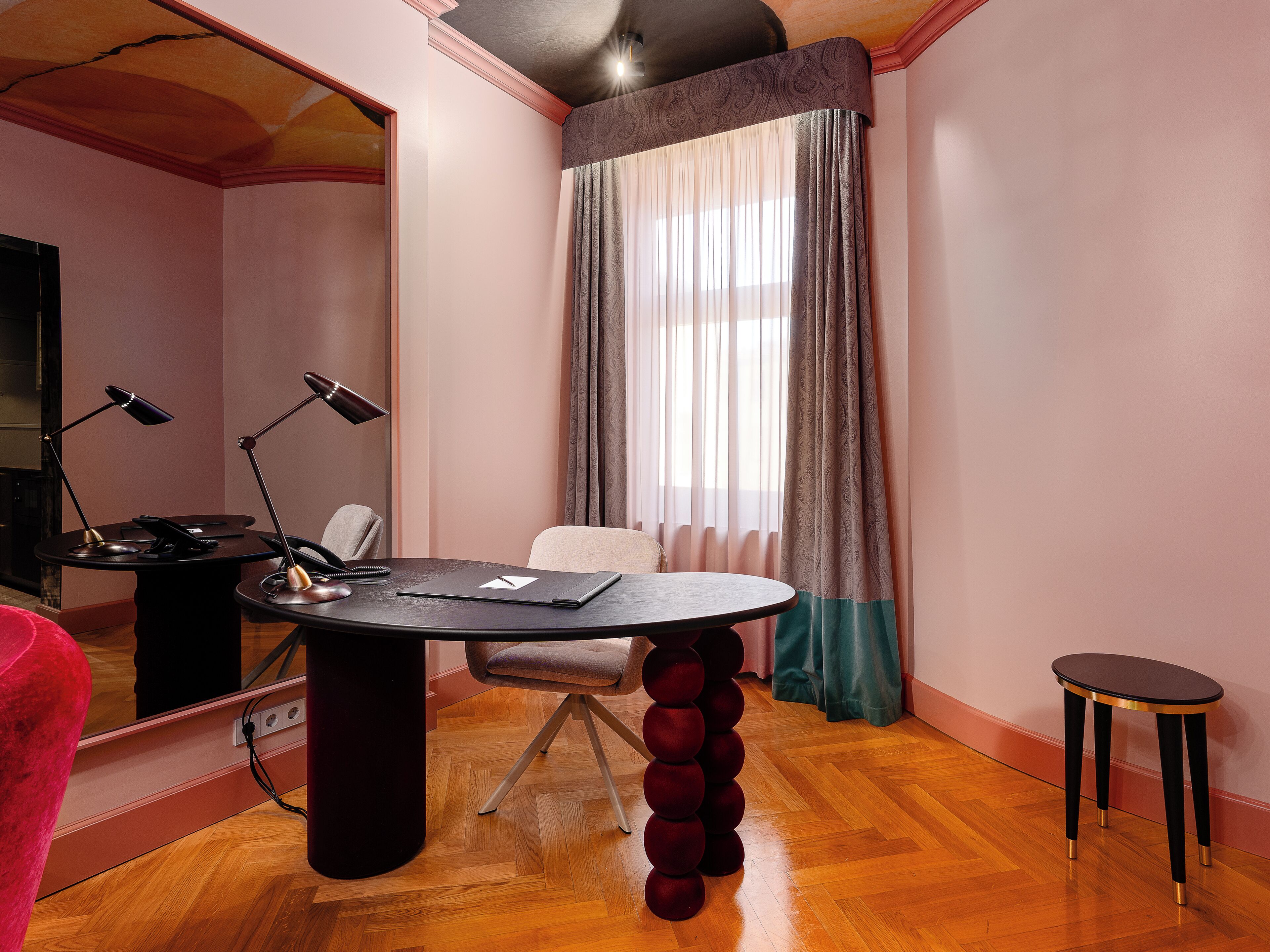 A room with a round table, a chair, a desk with a lamp, a TV, and a window with curtains.