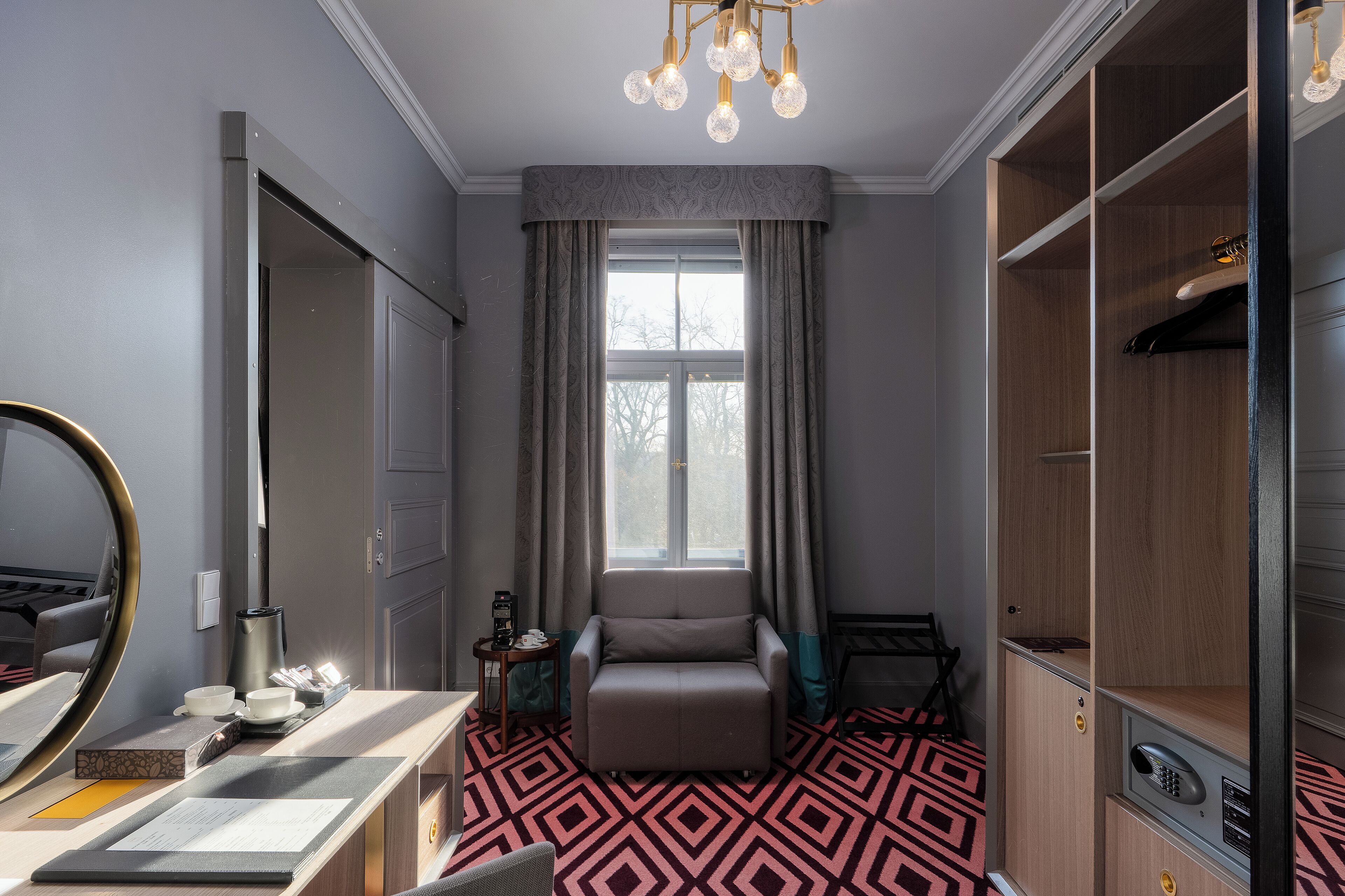 A compact hotel room with a bed, a vanity with a mirror, a small table with a kettle, and a window with curtains.