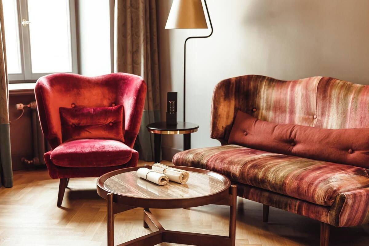 A room with a red velvet armchair, a patterned sofa, a small wooden coffee table with rolled-up papers, and a floor lamp.