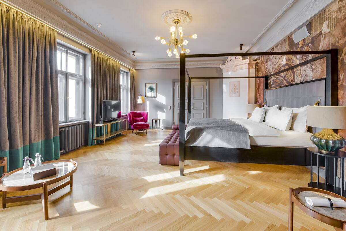 A spacious bedroom with a large bed, a sitting area with a pink chair, a television, and a dining table.