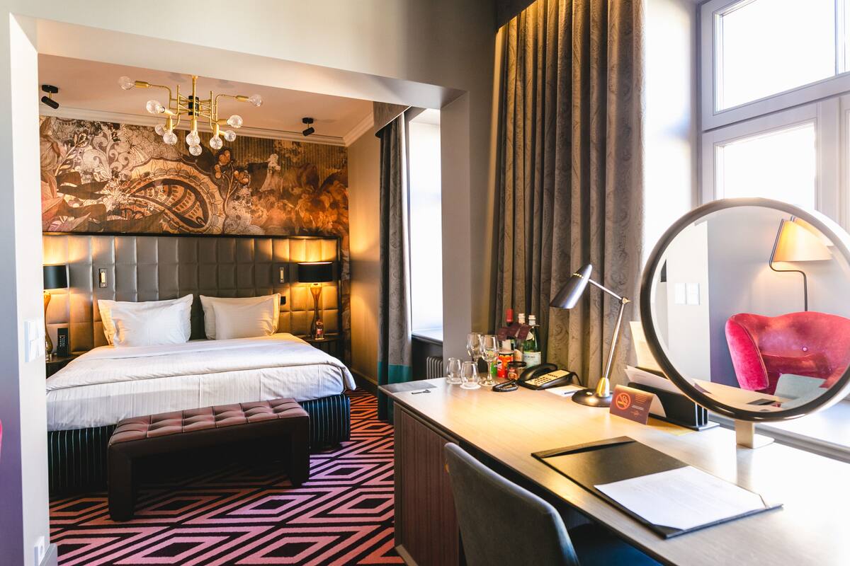 A hotel room with a large bed, a desk with a mirror, a red chair, and a wall with a decorative tapestry.