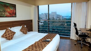 Habitación de hotel con una cama grande, un escritorio y vistas de la ciudad a través de un ventanal.