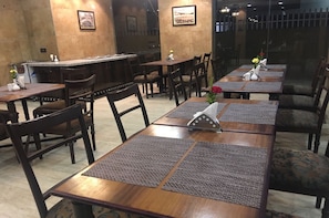 A restaurant with wooden tables and chairs, some with placemats. There are framed pictures on the walls and a bar area. - Hotel Mumbai House (Mumbai)