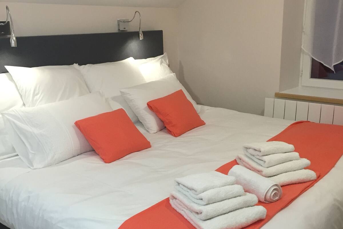 Un lit bien fait, avec une literie blanche et orange, une fenêtre avec des rideaux et un radiateur.