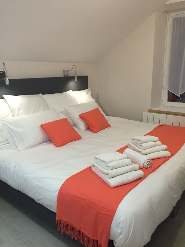 A neatly made bed with white and orange bedding, a window with curtains, and a radiator. - Hôtel le Bout du Monde (Saint-Georges)