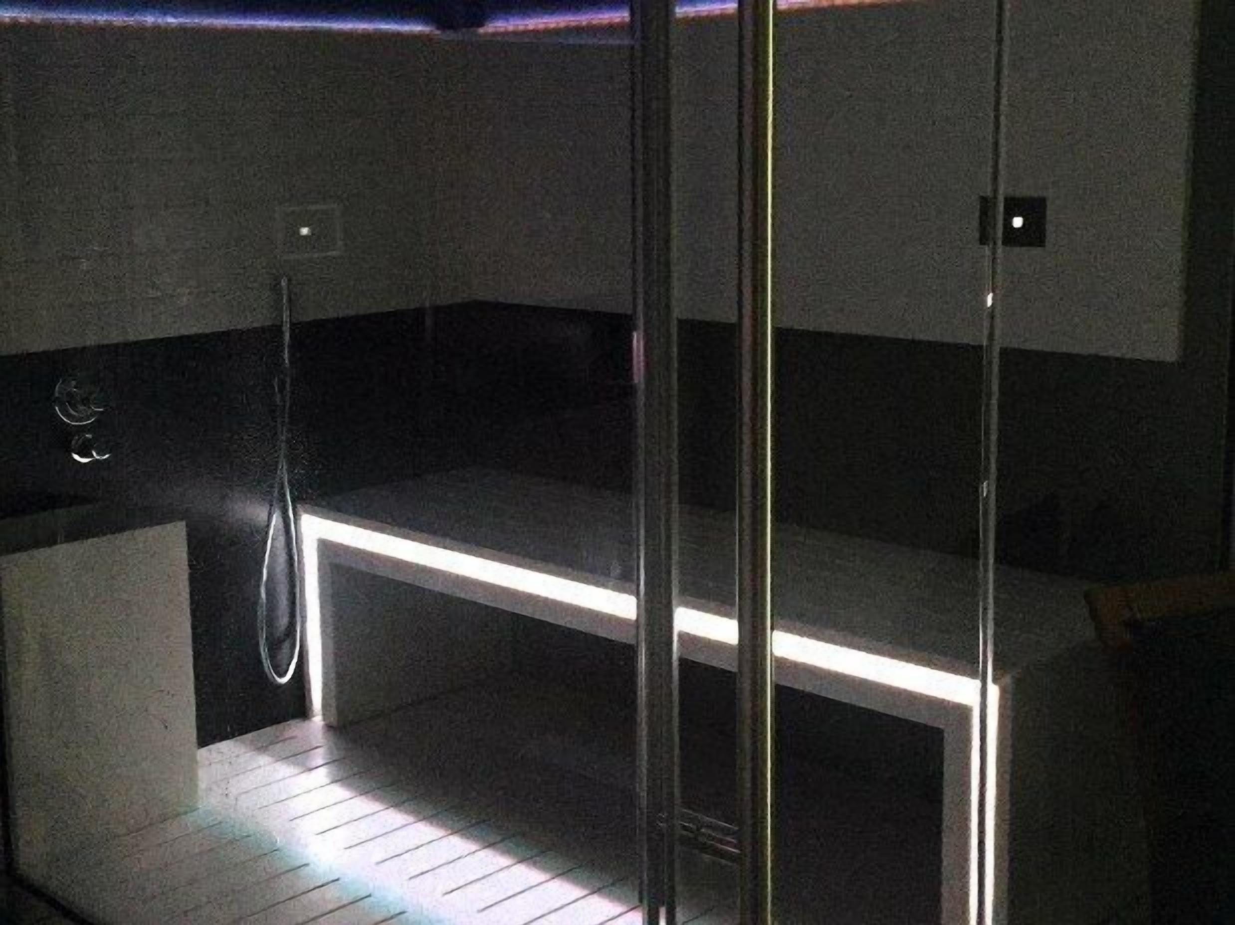 A modern, dimly lit sauna with a glass partition, a wooden bench, and a metal showerhead.