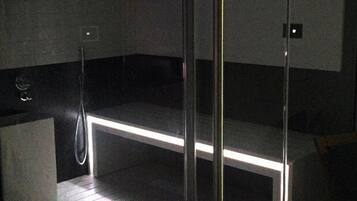 A modern, dimly lit sauna with a glass partition, a wooden bench, and a metal showerhead.